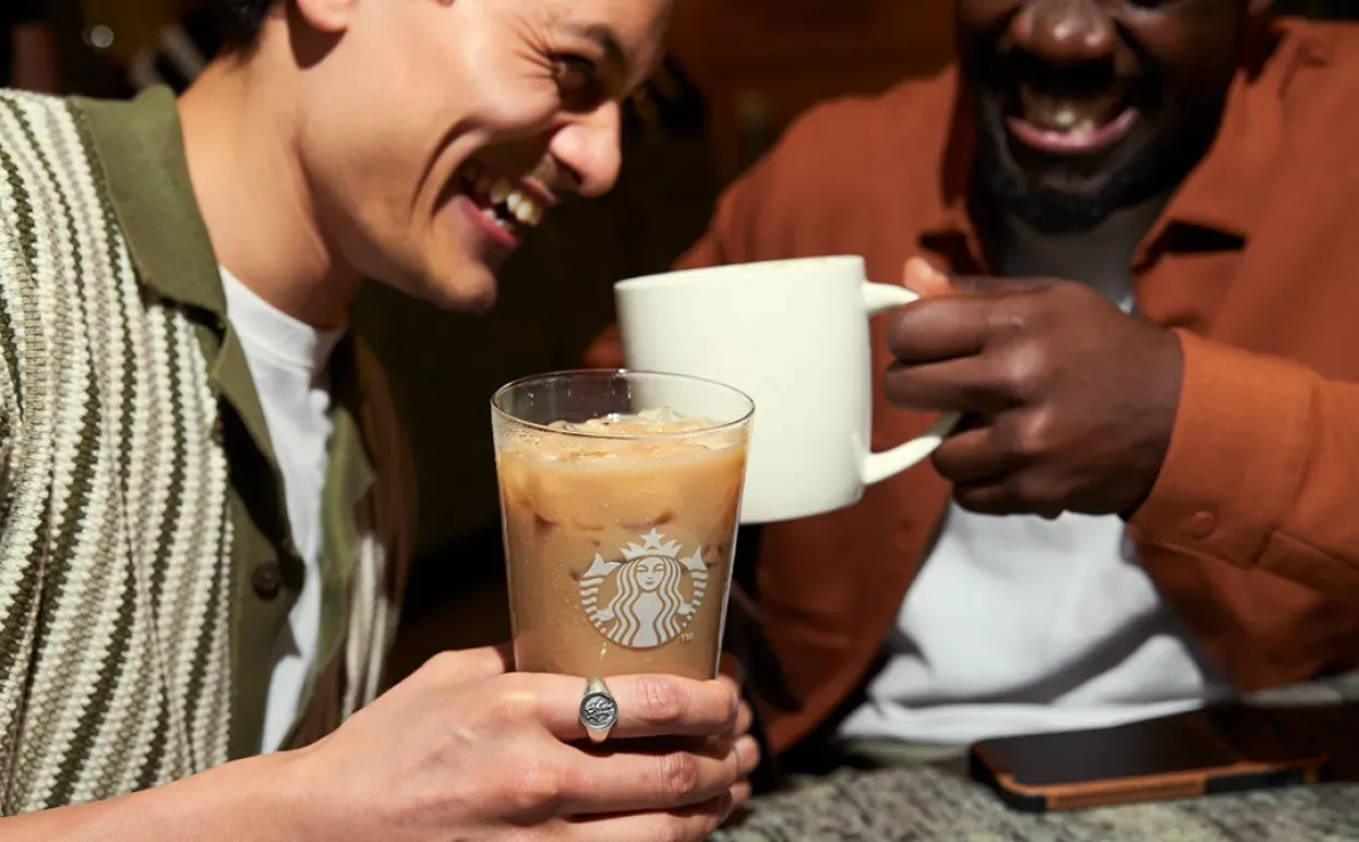 Dois clientes riem juntos, um segura uma bebida fresca num copo enquanto o outro tem uma bebida quente numa caneca clássica da Starbucks